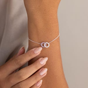 May include: A delicate silver chain bracelet featuring two interlocking circular charms. One circle is encrusted with small, clear white stones, while the other circle is set with small, light purple stones, creating a subtle contrast in color.