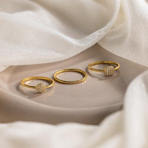May include: Three gold rings with small, round, and square-shaped stones set in them. The rings are on a light brown surface.