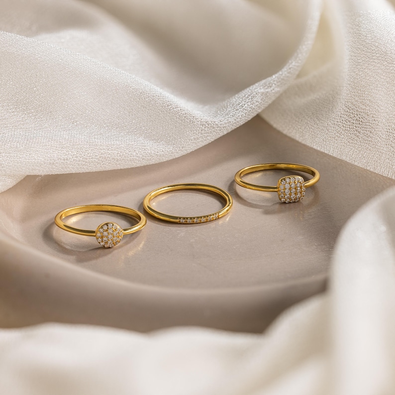 May include: Three gold rings with small, round, and square-shaped stones set in them. The rings are on a light brown surface.
