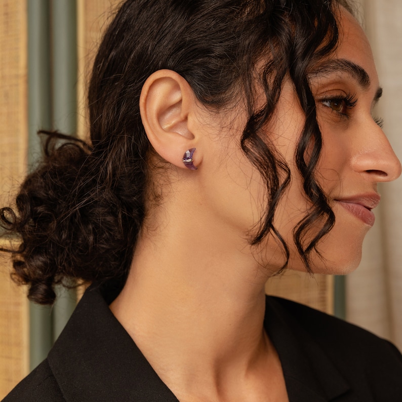 May include: A person wearing a pair of small, purple, crescent moon shaped earrings.