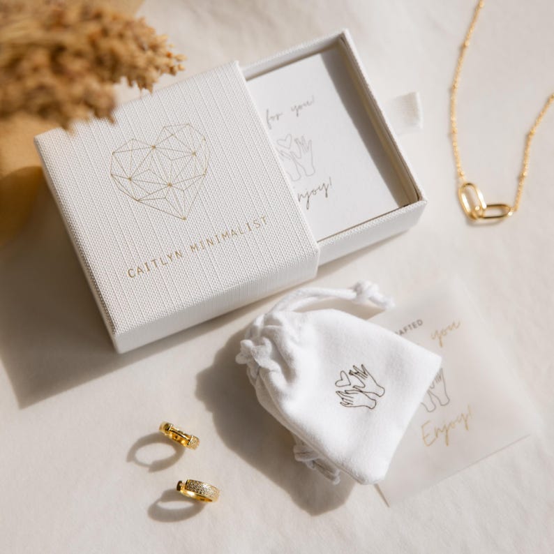 May include: A white jewelry box with the text "CAITLYN MINIMALIST" and a gold geometric heart design. A gold necklace and earrings are displayed with a small white bag and a card that says "for you" and "Enjoy!".