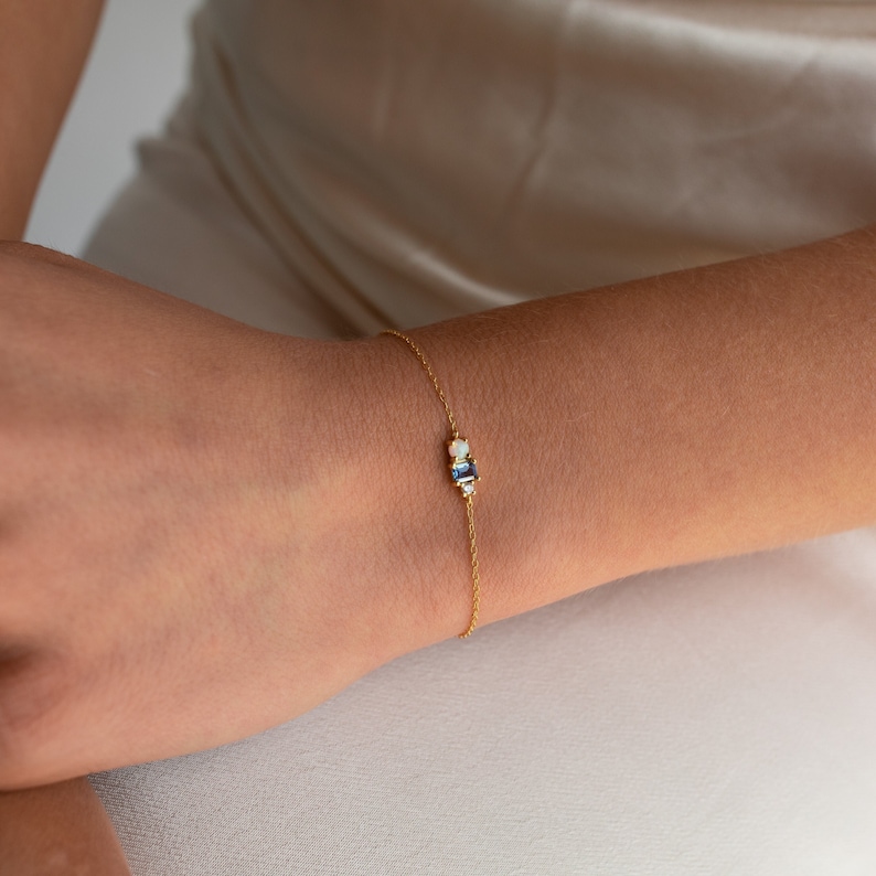 A close up of our Eloise Gemstone Bracelet in 18K Gold finish worn on our models wrist. Featuring a round opal stone next to a light blue rectangular turquoise stone, with a smaller clear CZ diamond stone at the end on a dainty cable chain.