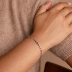 May include: A silver bracelet with a knot design. The bracelet is thin and delicate, and it is shown on a person's wrist.