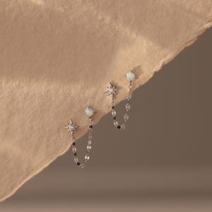 May include: A pair of silver-toned double-stud earrings connected by a delicate chain. Each earring features a small, round, iridescent opal-like stone and a four-pointed star charm with a tiny embedded clear stone, linked by a thin metal chain.