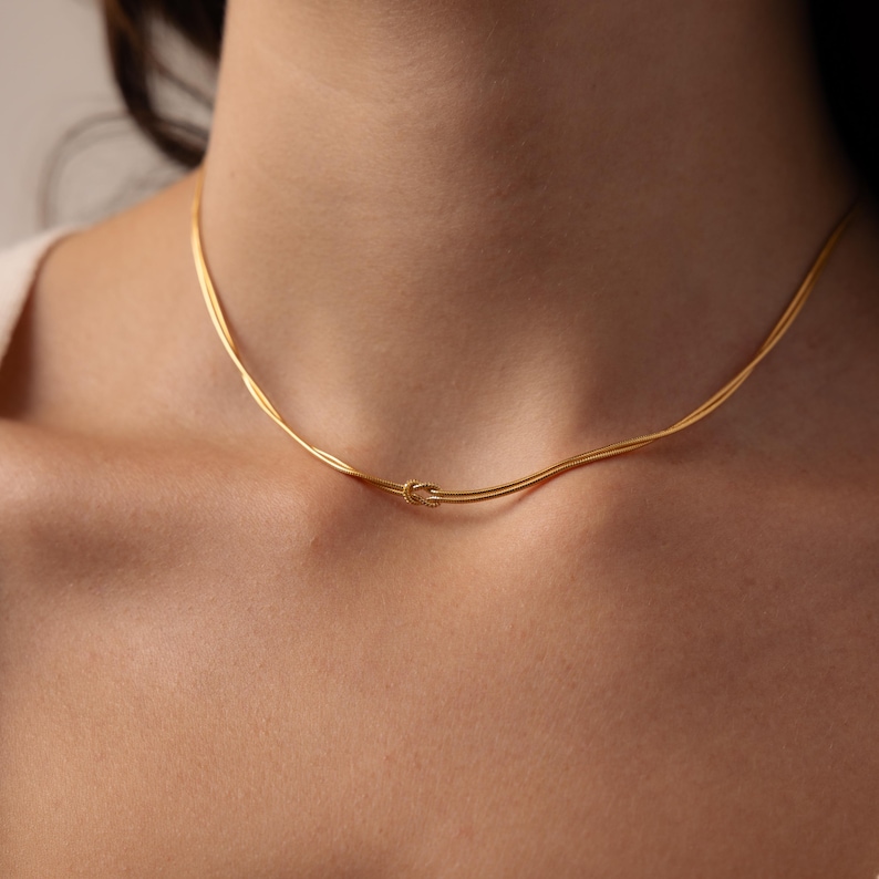 May include: A delicate gold necklace featuring a knot design. The necklace is made of thin, twisted strands of gold, creating a simple yet elegant look. The knot is centered, adding a focal point to the piece. The necklace is worn around the neck.