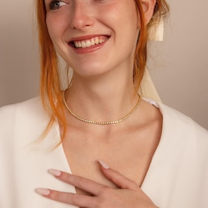 Puede incluir: Un collar de tenis en tono dorado que presenta una hilera continua de pequeñas piedras redondas y brillantes engastadas en una cadena metálica. El collar se ajusta al cuello, mostrando un diseño uniforme con acabado pulido y cierre.