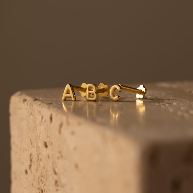 Three small gold initial stud earrings with the letters A, B, and C, displayed on a stone surface with a soft warm background.