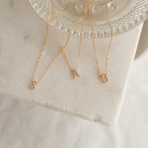 May include: Gold-toned necklaces with initial pendants. The pendants are encrusted with small, sparkling stones. The letters "S", "A", and "B" are visible. The necklaces are displayed on a white marble surface.