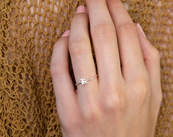May include: A delicate silver ring featuring the letter "B" in an elegant script. The ring is displayed on a hand with painted nails, set against a textured, golden-brown background. The ring is a minimalist piece of jewelry.