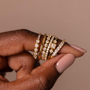 May include: A collection of gold rings with various designs, including bands with round and square-cut clear gemstones. The rings are stacked on a finger, showcasing different styles and textures. The background is a solid, neutral color.