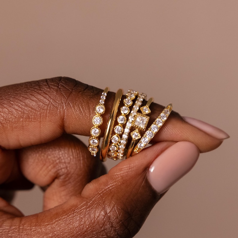 May include: A collection of gold rings with various designs, including bands with round and square-cut clear gemstones. The rings are stacked on a finger, showcasing different styles and textures. The background is a solid, neutral color.