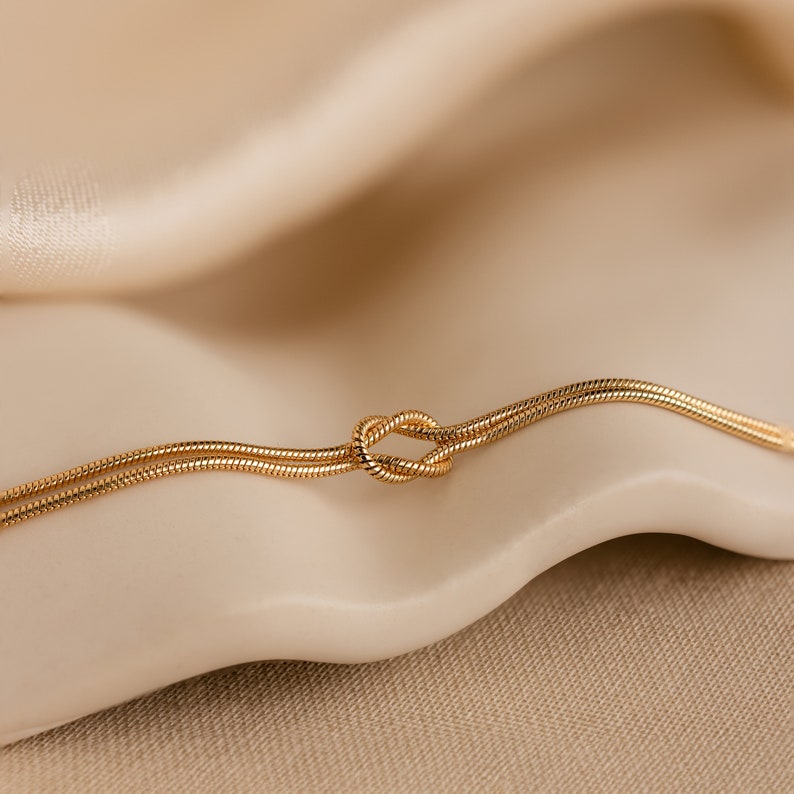 May include: A gold chain necklace with a knot design. The chain is thin and delicate, and the knot is a simple, elegant design.