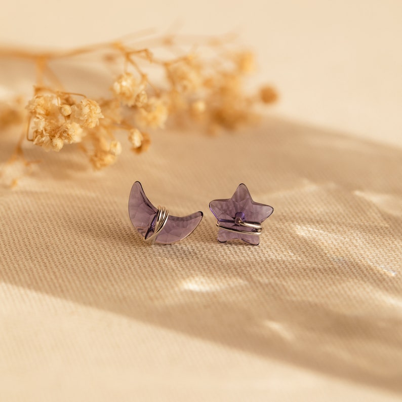 May include: A pair of earrings featuring a crescent moon and a star shape. Both are made of clear purple glass and wrapped in silver wire.