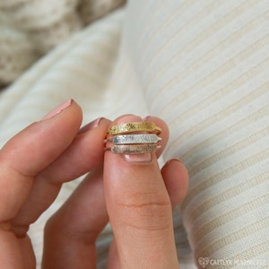 May include: Three fingerprint rings in gold, silver, and rose gold. Each ring features a detailed fingerprint design across the top. The rings are stacked, showcasing the different metal colors. The background is a soft, neutral color.