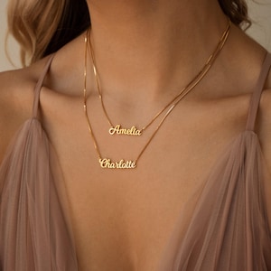 May include: Two gold-colored necklaces with delicate chains. The top necklace reads "Amelia" in cursive script, and the bottom necklace reads "Charlotte." The necklaces are layered, creating a stylish and personalized accessory.