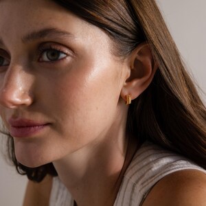 May include: Close-up of a person wearing small, gold hoop earrings. The earrings have a textured, beaded design. The person is wearing a striped, light-colored top.