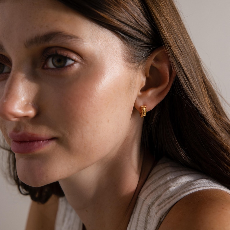 May include: Close-up of a person wearing small, gold hoop earrings. The earrings have a textured, beaded design. The person is wearing a striped, light-colored top.