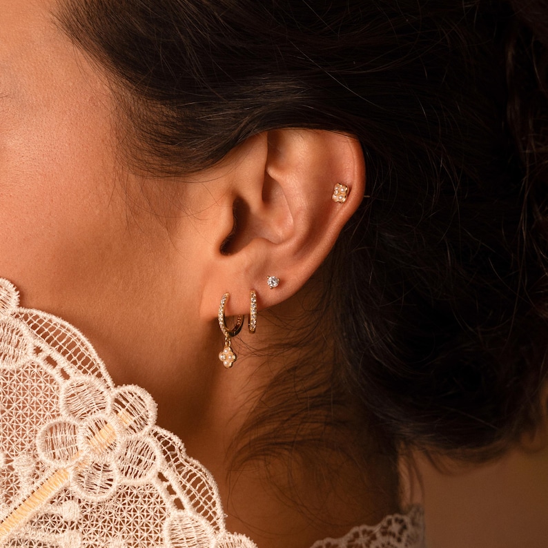 May include: Close-up of an ear adorned with multiple gold earrings. The ear features a small, square stud, a tiny round stud, and two hoop earrings. One hoop has a dangling charm, and the other is encrusted with small stones. The earrings are set against dark hair.