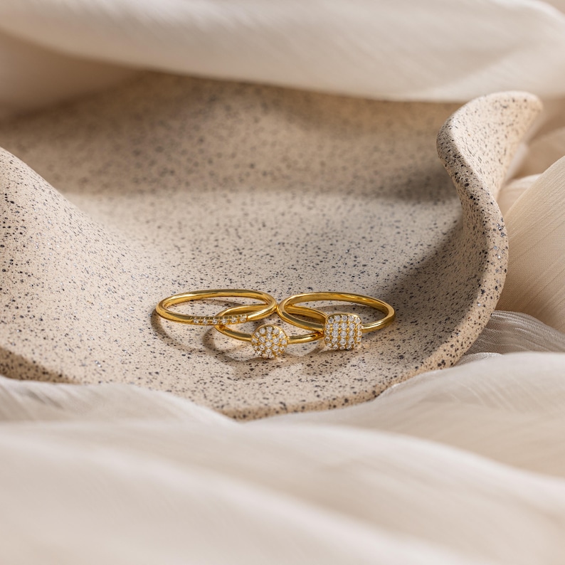 May include: Three gold rings with small, round, and square settings encrusted with tiny diamonds. The rings are displayed on a textured, light brown surface.