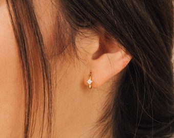 May include: A gold hoop earring with a small, square, clear gemstone. The earring is worn on the ear. The background is blurred, with dark hair visible.