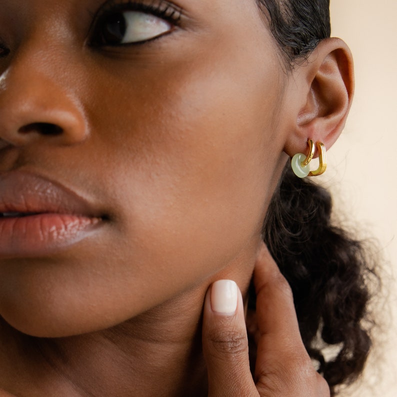 Peut inclure: Gros plan sur l'oreille d'une personne avec deux cr&eacute;oles en or. La cr&eacute;ole du haut a un charme en pierre vert clair.