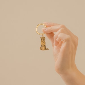 May include: A gold-colored metal keychain with a detailed engraved image of a dog. The dog is sitting with its head tilted up and its tail curled.