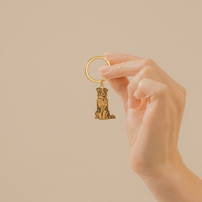 May include: A gold-colored metal keychain with a detailed engraved image of a dog. The dog is sitting with its head tilted up and its tail curled.
