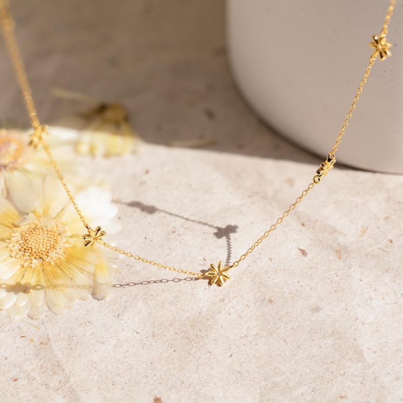 May include: A gold chain necklace with small flower charms. The necklace is on a light brown surface with dried flowers.