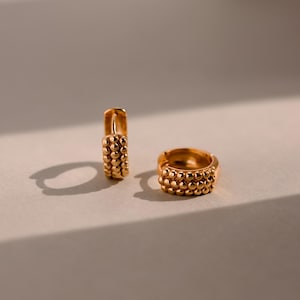 May include: Two gold-colored hoop earrings. Each earring features a textured design of small, evenly spaced spheres. The earrings are displayed on a neutral surface, with soft lighting creating shadows.