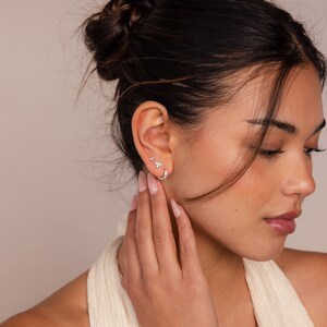 May include: A woman wearing a silver hoop earring and two silver stud earrings in her left ear. The earrings are small and delicate.