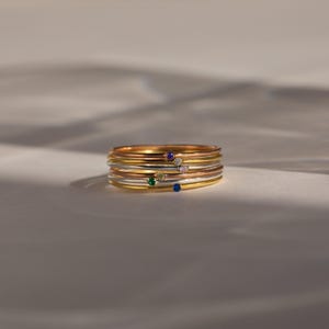 May include: A stack of six thin metal rings in gold, silver, and rose gold tones. Each ring features a single small, round, bezel-set gemstone in various colors, including blue, green, yellow, pink, white, and dark blue, arranged in a vertical line.