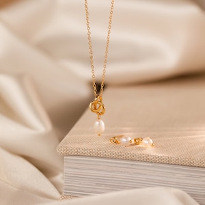 May include: A gold-colored necklace with a knot design and a dangling pearl, paired with matching pearl stud earrings. The jewelry set is displayed on a neutral-toned surface, creating an elegant and minimalist aesthetic.
