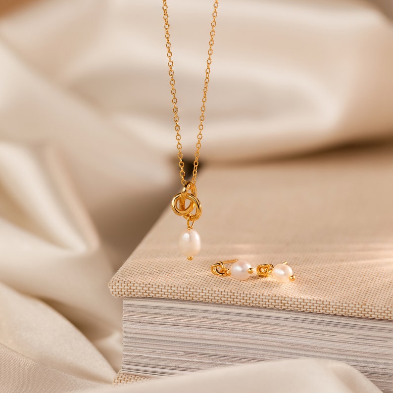 May include: A gold-colored necklace with a knot design and a dangling pearl, paired with matching pearl stud earrings. The jewelry set is displayed on a neutral-toned surface, creating an elegant and minimalist aesthetic.