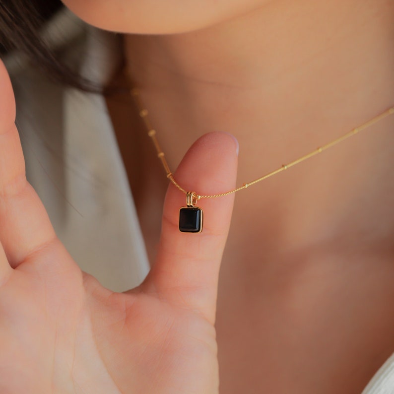 A close up of our Waverly Square Pendant Necklace in 18K Gold finish with the black charm held up by the models thumb  - featuring an about 8mm Black Enamel Square Pendant with a beaded satellite chain.