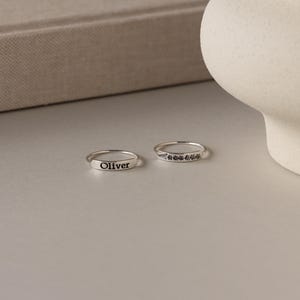 Puede incluir: Dos anillos de plata. Uno está grabado con el nombre "Oliver". El otro presenta un diseño de hojas. Los anillos están sobre una superficie gris claro.