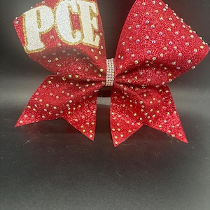 May include: A red glitter cheer bow featuring the letters PCE in white, textured block font on the left loop. The entire bow is embellished with scattered iridescent rhinestones and has a central band of small, clear, sparkling crystals.