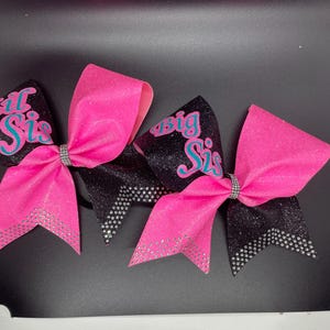 May include: Two pink and black cheer bows with glitter and rhinestones. The bows have the words "Big Sis" and "Lil Sis" written on them in blue.