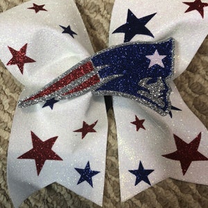 May include: White glitter hair bow with a red, white, and blue glitter football team logo and red, white, and blue glitter stars.