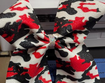 May include: A red, black, and white camouflage cheer bow with a sparkly finish and a rhinestone center.
