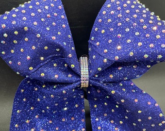 May include: A large, sparkly blue cheerleading bow. The bow is covered in iridescent rhinestones and glitter. A silver band with more rhinestones secures the center of the bow. The bow is a vibrant accessory.