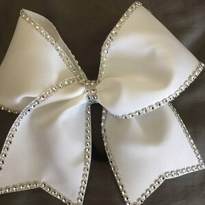 Pu&ograve; includere: Un fiocco da cheerleader bianco con un bordo di strass argentati. Il fiocco &egrave; legato in una forma grande e simmetrica.