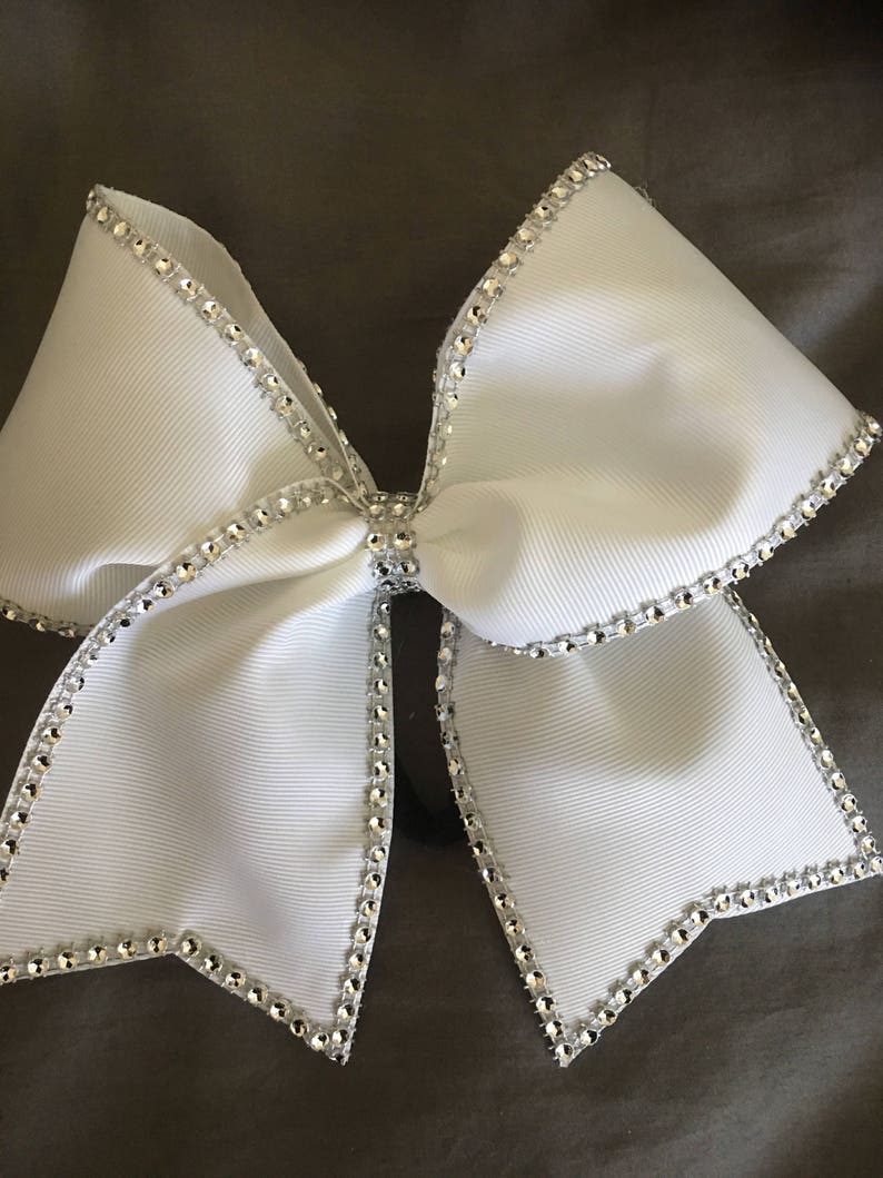 Pu&ograve; includere: Un fiocco da cheerleader bianco con un bordo di strass argentati. Il fiocco &egrave; legato in una forma grande e simmetrica.