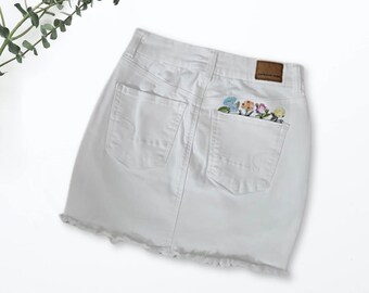 May include: White denim mini skirt with a frayed hem and a small embroidered flower patch on the back pocket. The patch features a variety of colorful flowers. The label "American Eagle" is visible on the back pocket.