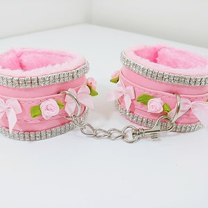Op de afbeelding: Een paar roze nep bont handboeien met een zilveren ketting en strassversieringen. Elke boeien heeft een roze satijnen strik en een kleine roze roos.
