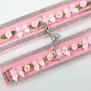 Op de afbeelding: Een paar roze nep bontmanchetten met zilveren strass steentjes en roze bloemen van stof. De manchetten zijn verbonden door een zilveren ketting.