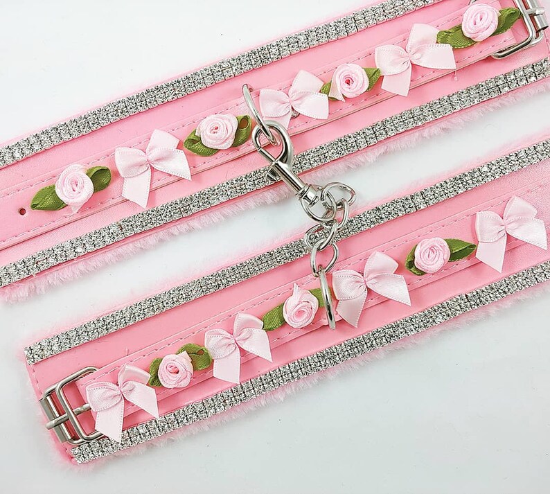 Op de afbeelding: Een paar roze nep bontmanchetten met zilveren strass steentjes en roze bloemen van stof. De manchetten zijn verbonden door een zilveren ketting.