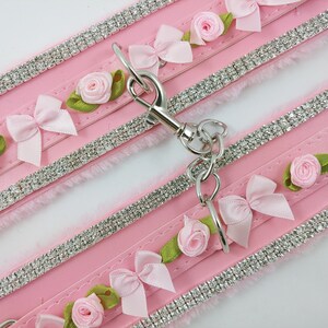 Op de afbeelding: Twee roze kunstbont halsbanden met rijen strass steentjes en roze strikken en rozen. De halsbanden zijn verbonden door een zilveren ketting met een sluiting.