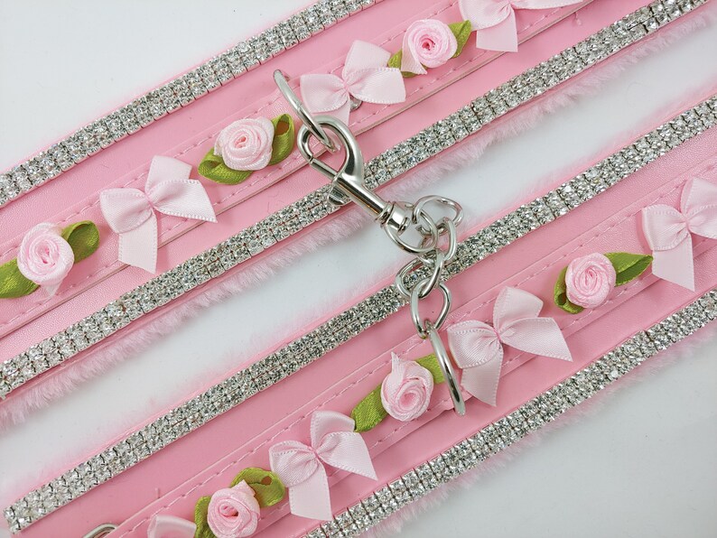 Op de afbeelding: Twee roze kunstbont halsbanden met rijen strass steentjes en roze strikken en rozen. De halsbanden zijn verbonden door een zilveren ketting met een sluiting.