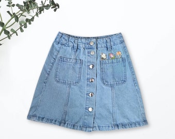 May include: A light blue denim skirt with a button front closure and two front pockets. The skirt has embroidered flowers on the right pocket.