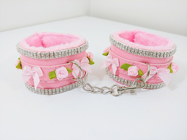 Op de afbeelding: Een paar roze nep bont handboeien met een zilveren ketting en strassversieringen. Elke boeien heeft een roze satijnen strik en een kleine roze roos.